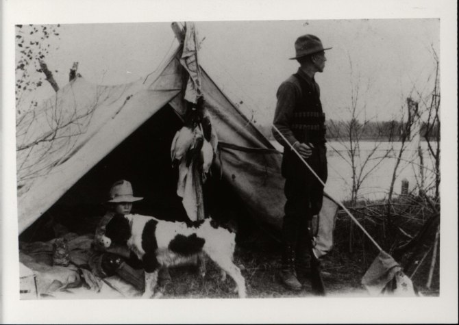 Black and white image of two men and their dog near a tent