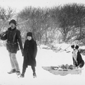 Black and white winter image of two kids standing with a dog sitting a sled