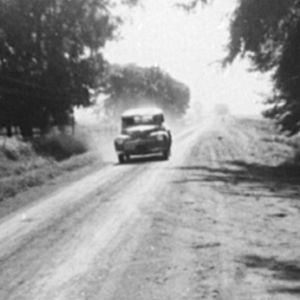 Black and white photo of older vehicle driving down a tree-lined country road churning up dust in the background.